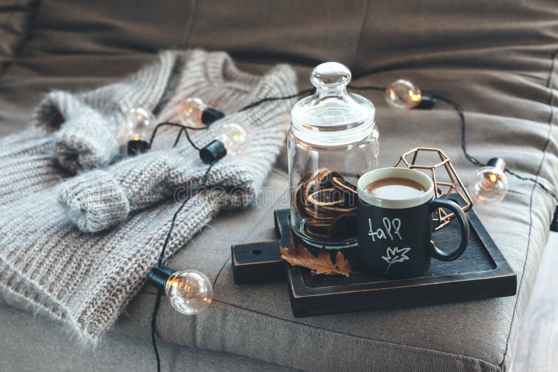 Still life details of living room. Cup of coffee on rustic wooden tray, candle and warm woolen sweater on sofa, decorated with led lights. Autumn weekend concept. Fall home decoration. Still life details of living room. Cup of coffee on rustic wooden tray, candle and warm woolen sweater on sofa, decorated with led lights. Autumn weekend concept. Fall home decoration.