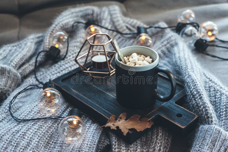 Still life details of living room. Cup of coffee on rustic wooden tray, candle and warm woolen sweater on sofa, decorated with led lights. Autumn weekend concept. Fall home decoration. Still life details of living room. Cup of coffee on rustic wooden tray, candle and warm woolen sweater on sofa, decorated with led lights. Autumn weekend concept. Fall home decoration.