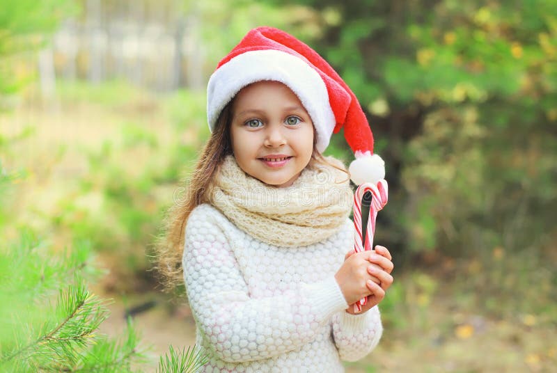 Concetto di Natale - bambino della bambina del ritratto in cappello rosso di Santa con la canna dolce della lecca-lecca immagini stock Concetto di Natale - bambino della bambina del ritratto in cappello rosso di Santa con la canna dolce della lecca-lecca immagini stock