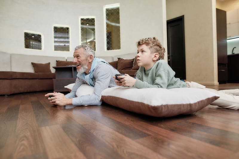Concorso online. vista laterale di un nonno con un nipote sdraiato sui cuscini per terra e che gioca ai videogiochi immagine stock