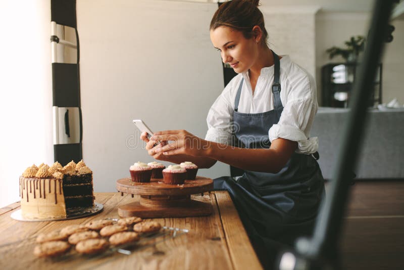 Young woman taking pictures of a cake in the kitchen. Female baker capturing photos of pastry items with her mobile phone. Young woman taking pictures of a cake in the kitchen. Female baker capturing photos of pastry items with her mobile phone