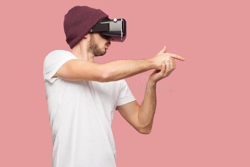 Confident bearded young hipster man in white shirt and casual hat standing, wearing vr playing video game, showing gun sing with stock image Confident bearded young hipster man in white shirt and casual hat standing, wearing vr playing video game, showing gun sing with stock image