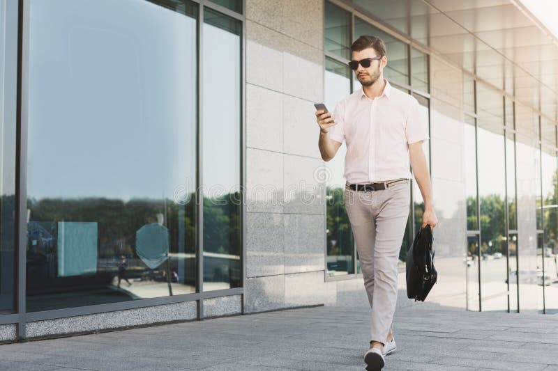 Confident successful businessman using smartphone outdoors. Young executive checking email on phone, walking at modern building, copy space. Confident successful businessman using smartphone outdoors. Young executive checking email on phone, walking at modern building, copy space