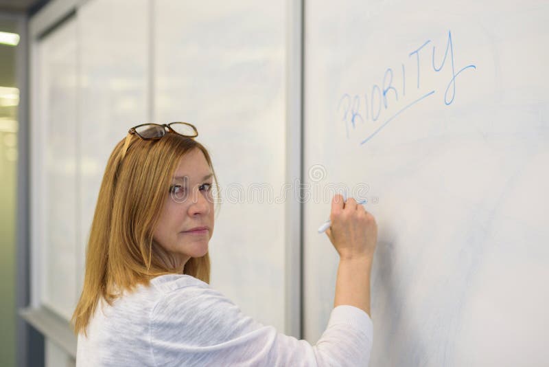 Confident businesswoman writing word priority on whiteboard in m