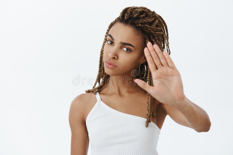 Confident serious-looking displeased and annoyed african american woman with dreadlocks raising palms to decline and say no demanding person stop shooting being irritated over gray wall.