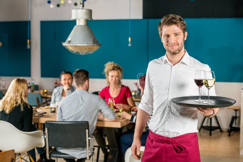Confident Waiter Holding Tray At Restaurant. Portrait of confident waiter holding tray at restaurant with customers in background stock photo Confident Waiter Holding Tray At Restaurant. Portrait of confident waiter holding tray at restaurant with customers in background stock photo