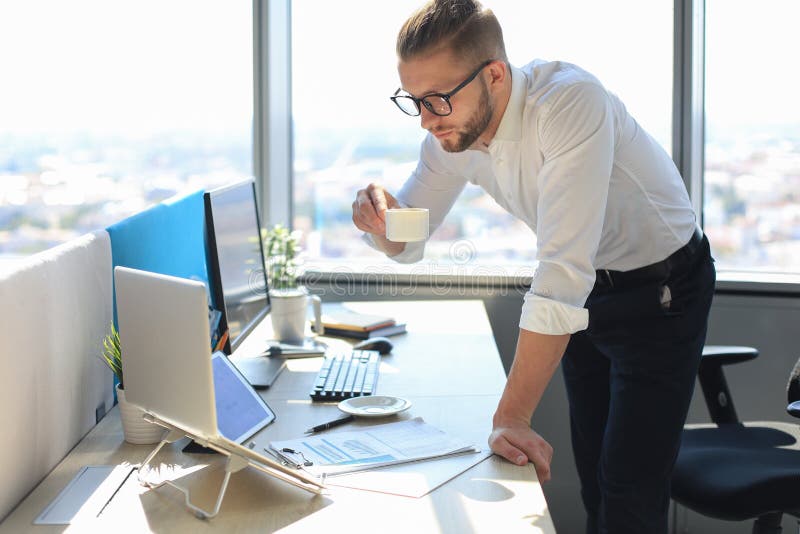 Confident young business man working on laptop and drinking from coffee cup while sitting in office royalty free stock photography