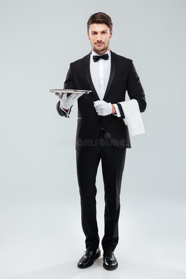 Confident young waiter in tuxedo standing and holding tray