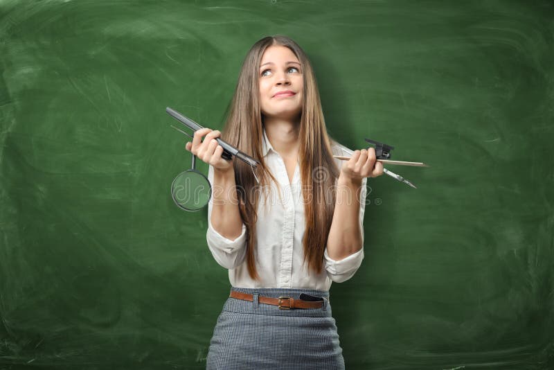 Confused businesswoman on green chalkboard background holding pens, ruler and magnifying glass in her hands. royalty free stock image