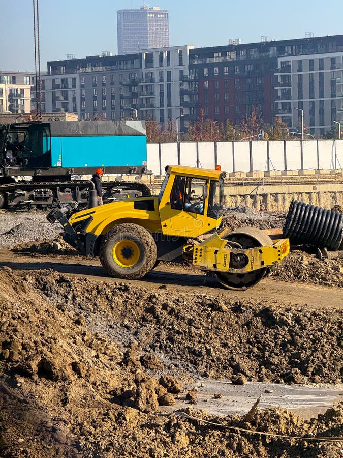 Construction roller moving across soil mound near residential buildings. Infrastructure development, engineering groundwork, roadwork phase and structural compaction activity. Load structural stock images, royalty-free photos and pictures