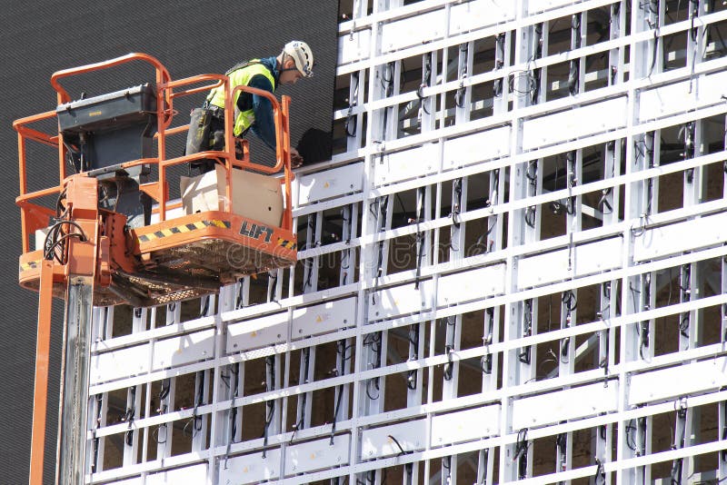 Construction worker in crane basket installing sheets cladding for covering structure on a building facade wall