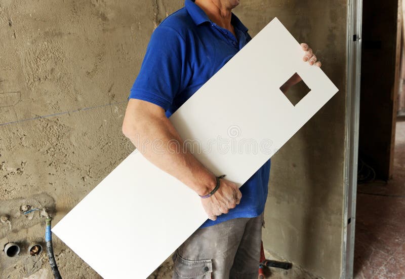Porcelain stoneware tiles for tiling of the kitchen with a square hole planned for electrical outlets installation. Male construction worker in a blue shirt holds a white porcelain stoneware tile featuring a square cutout. The background shows an unfinished plastered wall, with visible pipes and fittings. The tile is positioned vertically with the square hole intended for electrical outlets. The worker's posture suggests he is in the process of installation or preparation for tiling. Dust and construction materials are evident in the surroundings, indicating an active work area. Bricklayer tiler stock images, royalty-free photos and pictures