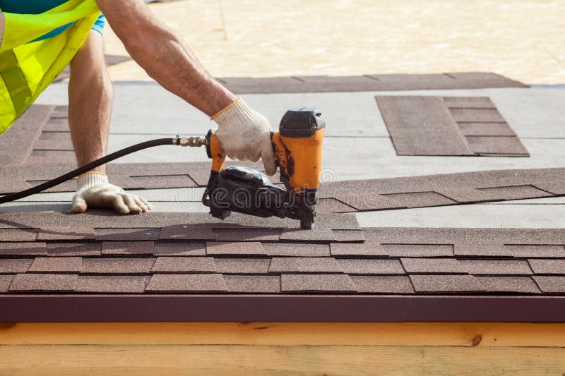 Construction worker putting the asphalt roofing shingles with nail gun on a new frame house.