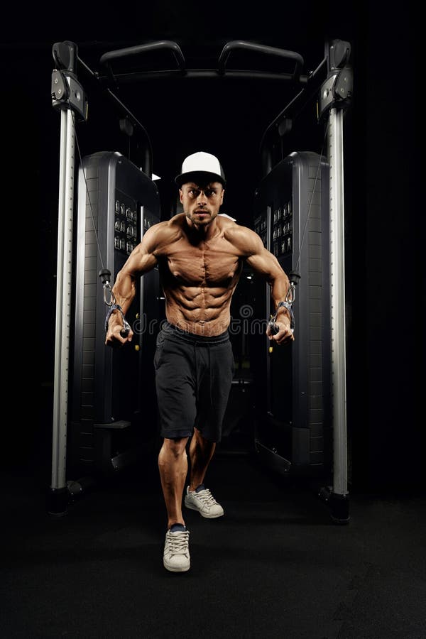 Constructor de carrocería masculino haciendo ejercicio de cruce de cables alto. Vista frontal del cofre de entrenamiento masculino muscular sin camisa en el gimnasio en atmósfera oscura Hombre guapo con cuerpo perfecto haciendo ejercicio de cruce de cables alto Concepto de deporte, construcción de cuerpos
