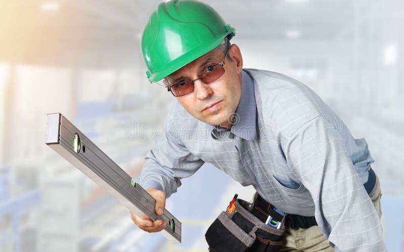 Builder with tools and in hard hat on head and with level in hands looks into camera with serious view. Builder with tools and in hard hat on head and with level in hands looks into camera with serious view