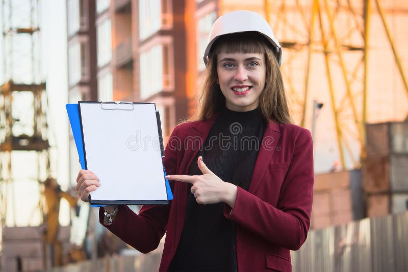 Construtor da mulher que guarda modelos, prancheta Menina de sorriso do arquiteto no capacete no fundo da construção fotografia de stock Construtor da mulher que guarda modelos, prancheta Menina de sorriso do arquiteto no capacete no fundo da construção fotografia de stock