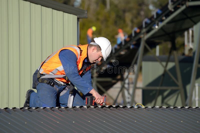 A builder wearing a safety harness for working at heights unscrews roofing iron on a building marked for demolition. A builder wearing a safety harness for working at heights unscrews roofing iron on a building marked for demolition