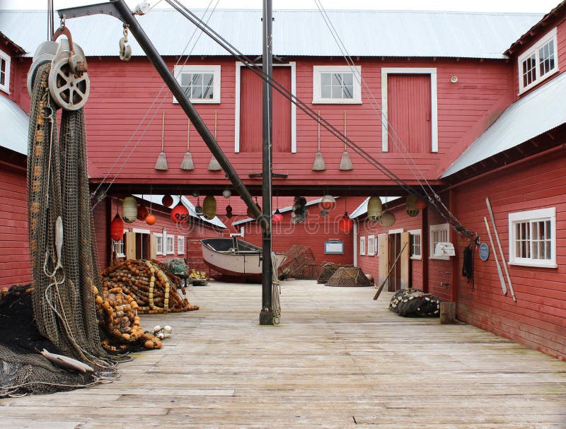 A red building is situated on a wharf. There is a fishing net draped over a roller on the left and a small white fishing boat in the back. A red building is situated on a wharf. There is a fishing net draped over a roller on the left and a small white fishing boat in the back.
