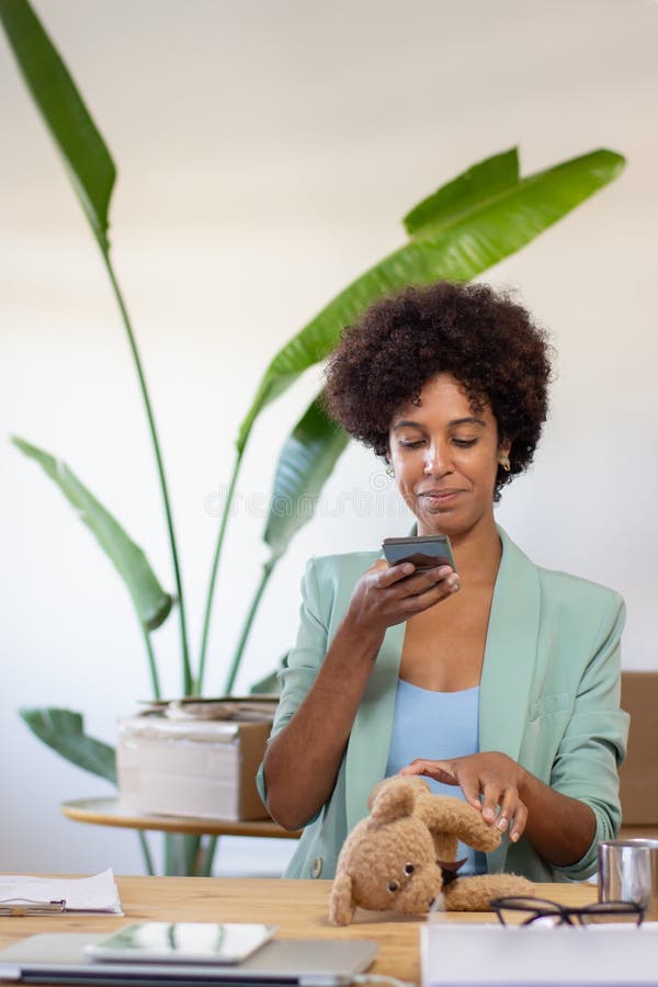 Content blackwoman photographing product in office. Attractive manager sitting at table and sending photo of toy to customer. Postal service concept