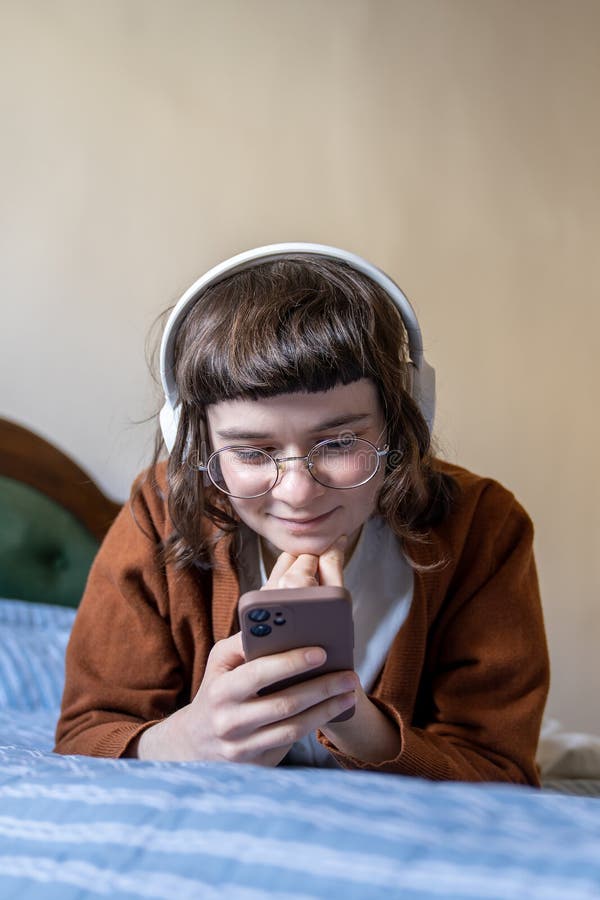 Contente jeune fille avec casque sans fil écouter de la musique vidéo appel avec des amis au téléphone dans le lit image stock