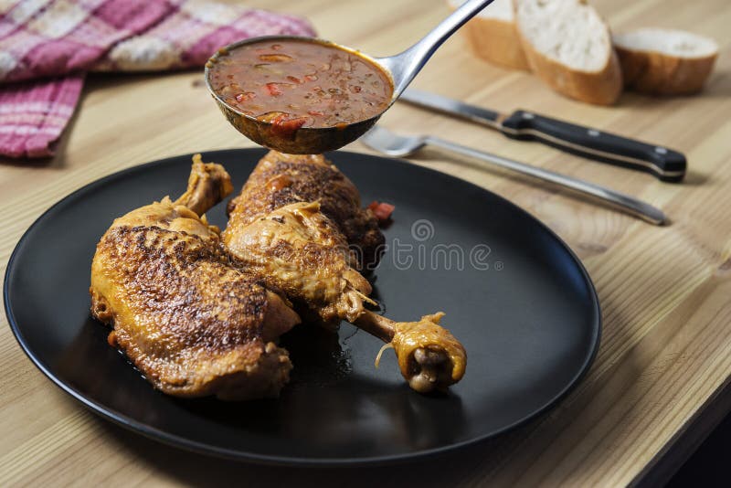 Ladle serving vegetable garnish in sauce over the chicken on the plate. On the wooden table there are cutlery, bread and a vintage napkin. Ladle serving vegetable garnish in sauce over the chicken on the plate. On the wooden table there are cutlery, bread and a vintage napkin