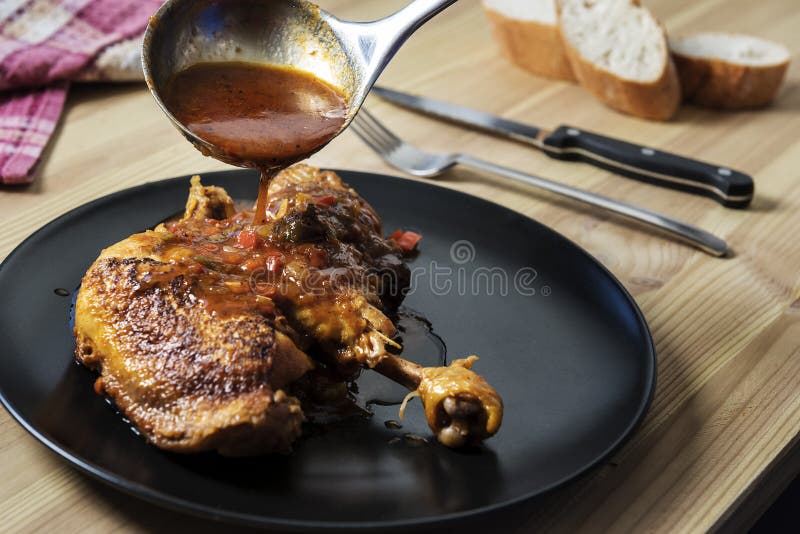 Ladle pouring vegetable garnish in sauce over the chicken on the plate. On the wooden table there are cutlery, bread and a vintage kitchen rag. Ladle pouring vegetable garnish in sauce over the chicken on the plate. On the wooden table there are cutlery, bread and a vintage kitchen rag