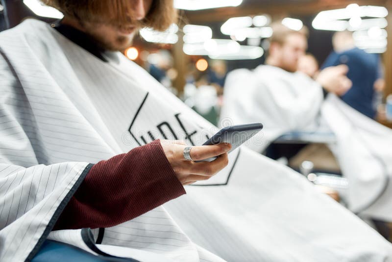 Conversando com um amigo. foto recortada de um jovem usando celular durante um corte de cabelo na barbearia ou no salão imagem de stock