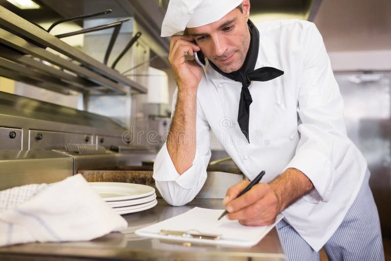 Cook Writing on Clipboard while Using Cellphone in Kitchen Stock Image
