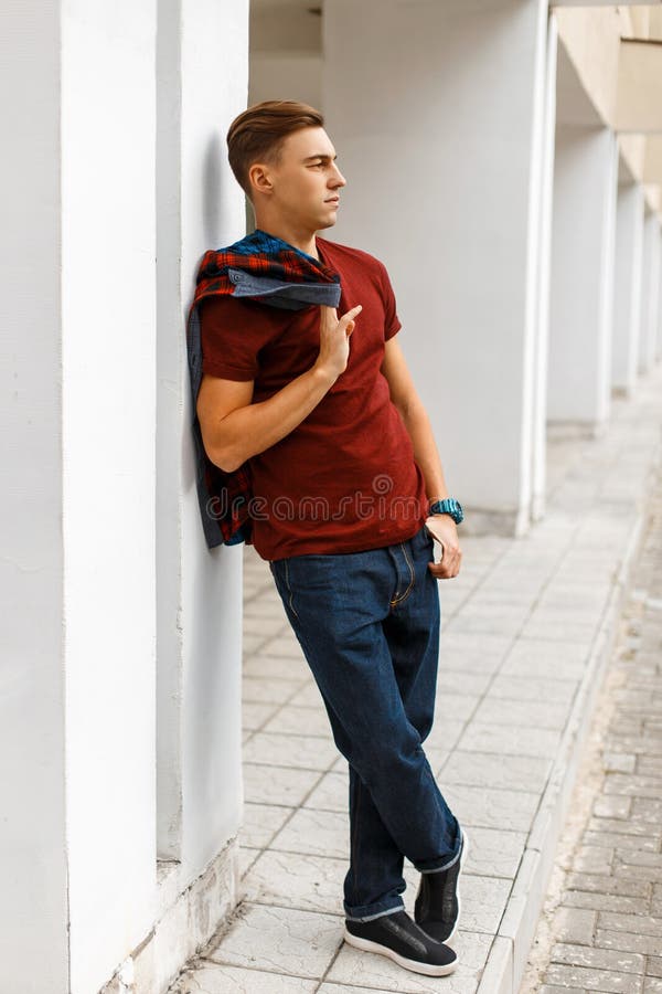 Cool handsome young man in trendy red t-shirt in a plaid fashionable shirt in blue jeans in sneakers posing on the street royalty free stock photography Cool handsome young man in trendy red t-shirt in a plaid fashionable shirt in blue jeans in sneakers posing on the street royalty free stock photography