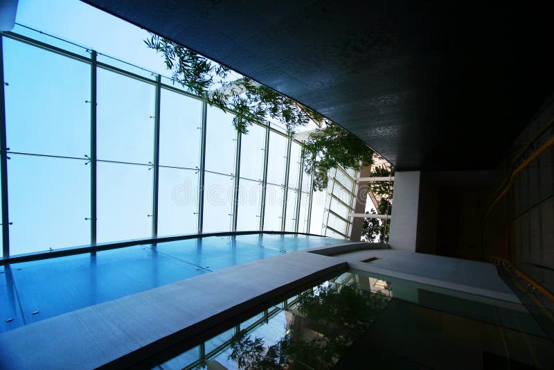 An image of the cool blue interior of a modern style hotel in contemporary architecture. The skylight above the corridor lets in sunlight, and conserves electrical energy in the day. Man-made waterfall over smooth black wall on the right, with plantings of bamboo trees on the roof top garden to complete the landscaping. Use of glass and steel gives an ultra modern look with clean lines and simple elegance. Man made waterfall stock images, royalty-free photos and pictures
