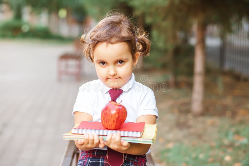 Copil mic și drăguț din școală în uniformă în școală în apropiere și ținând cărți de notițe și măr roșu stock images