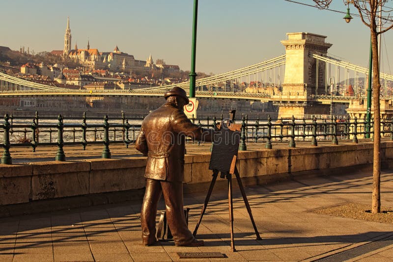 Budapest, Hungary-January 08, 2017:Copper statue of the painter who paints the landscape of the city of Budapest. Chain Bridge across River Danube and Medieval Fisherman`s Bastion in the background