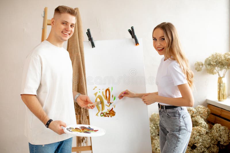 Coppia sorridente dipinge su tela usando spazzole a colori d'acqua vicino a una scala di legno e un mazzo di fiori secchi. Coppia di giovani felici e sorridenti dipingono su tela usando pennelli a colori d'acqua che si posano vicino a una scala di legno con un sacchetto di fiori secchi. workshop di pittura per terapie d'arte.