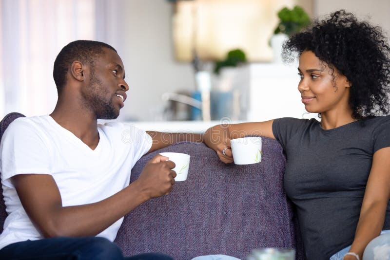 Satisfied African American couple talking, drinking coffee at home, smiling men and women sitting on cozy sofa at home, having pleasant conversation, spending weekend together, resting. Satisfied African American couple talking, drinking coffee at home, smiling men and women sitting on cozy sofa at home, having pleasant conversation, spending weekend together, resting