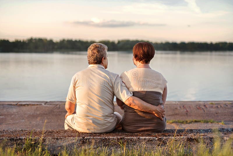 Coppie anziane della famiglia che si siedono sulla riva del lago e del fiume che guardano il tramonto immagine stock libera da diritti Coppie anziane della famiglia che si siedono sulla riva del lago e del fiume che guardano il tramonto immagine stock libera da diritti