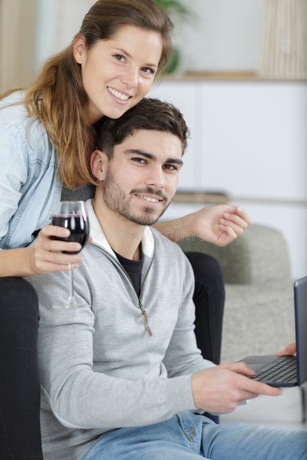 Coppie felici che si rilassano con il vino rosso ed il computer portatile in salone immagine stock libera da diritti Coppie felici che si rilassano con il vino rosso ed il computer portatile in salone immagine stock libera da diritti