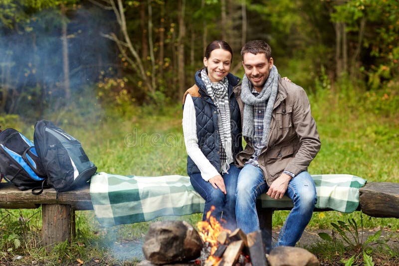 Camping, travel, tourism, hike and people concept - happy couple sitting on bench and warming near campfire at camp in woods. Camping, travel, tourism, hike and people concept - happy couple sitting on bench and warming near campfire at camp in woods