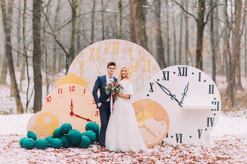 Coppie felici di nozze che posano ai grandi orologi d'annata nelle decorazioni creative della foresta di autunno fotografia stock Coppie felici di nozze che posano ai grandi orologi d'annata nelle decorazioni creative della foresta di autunno fotografia stock