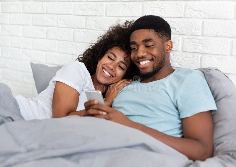 Coppie millenarie nere sorridenti facendo uso dello smartphone sul letto immagine stock libera da diritti Coppie millenarie nere sorridenti facendo uso dello smartphone sul letto immagine stock libera da diritti
