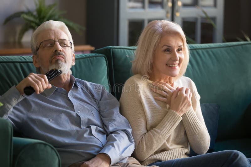 Aged couple, mature wife and husband watching tv show, series, drama or melodrama movie together at home, senior family enjoying weekend, bored men and smiling fascinated women sitting on couch. Aged couple, mature wife and husband watching tv show, series, drama or melodrama movie together at home, senior family enjoying weekend, bored men and smiling fascinated women sitting on couch