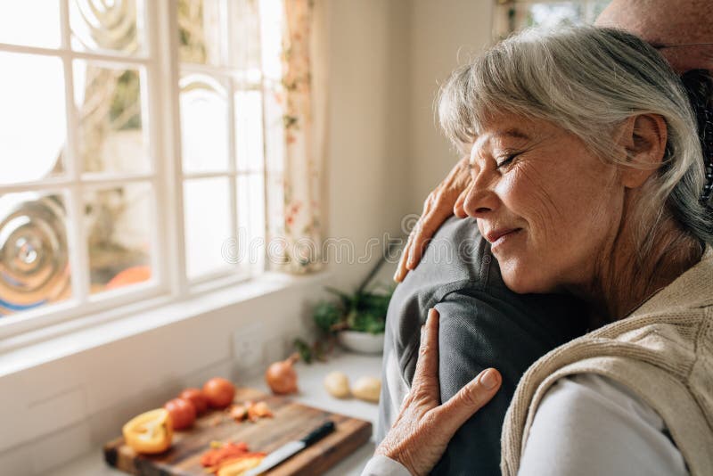 Coppie senior a casa che si abbracciano che sta nella cucina Donna senior che dà un abbraccio caldo al suo marito nell'espression fotografia stock libera da diritti Coppie senior a casa che si abbracciano che sta nella cucina Donna senior che dà un abbraccio caldo al suo marito nell'espression fotografia stock libera da diritti