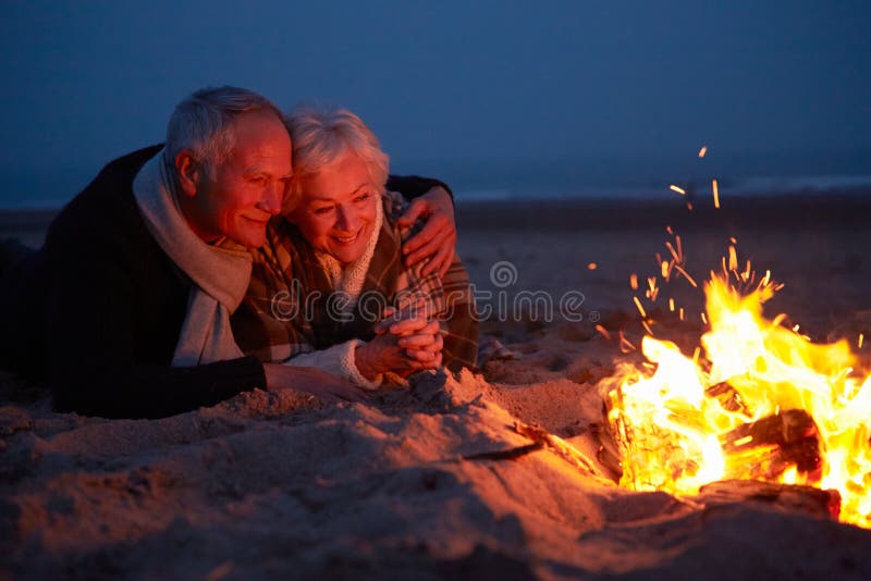 Coppie senior che si siedono dal fuoco sulla spiaggia di inverno fotografia stock Coppie senior che si siedono dal fuoco sulla spiaggia di inverno fotografia stock