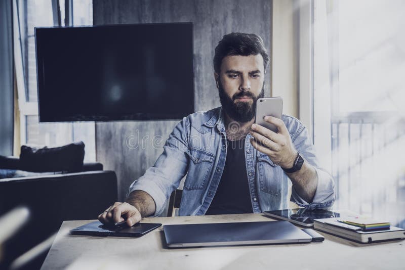 Young man sitting at window and working on laptop. Bearded man sitting at  desktop with smartphone in his hands. Online education. Stock Photo