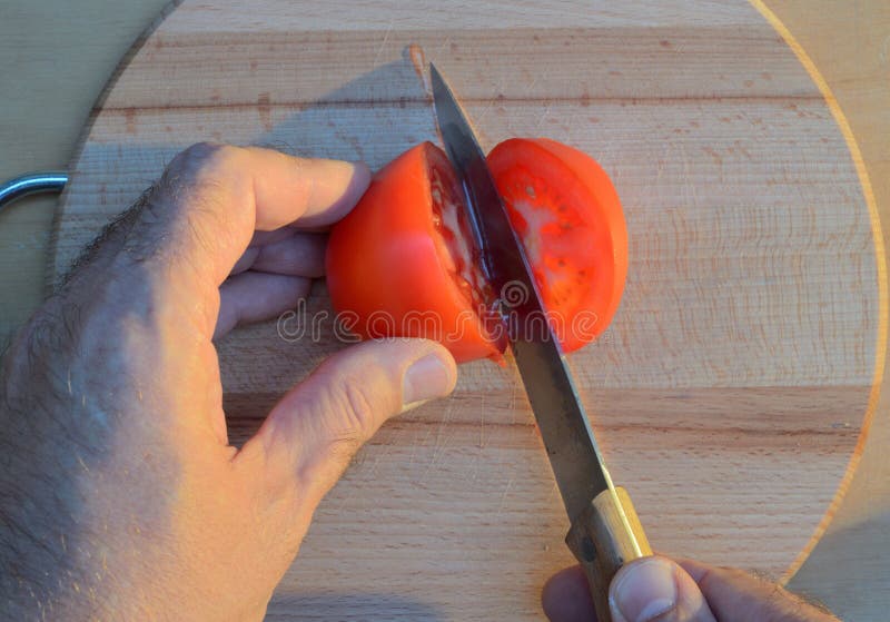 Cortar un tomate con las manos con un cuchillo en una tabla de cortar de madera en la puesta del sol fotos de archivo