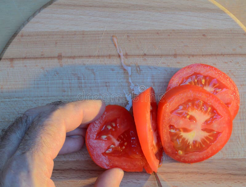 Cortar un tomate con las manos con un cuchillo en una tabla de cortar de madera en la puesta del sol fotos de archivo