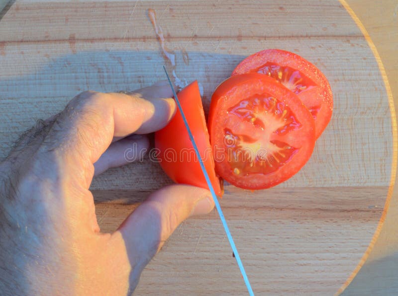 Cortar un tomate con las manos con un cuchillo en una tabla de cortar de madera en la puesta del sol fotos de archivo libres de regalías