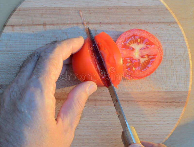 Cortar un tomate con las manos con un cuchillo en una tabla de cortar de madera en la puesta del sol foto de archivo libre de regalías