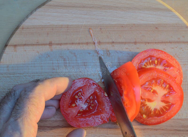 Cortar un tomate con las manos con un cuchillo en una tabla de cortar de madera en la puesta del sol fotografía de archivo