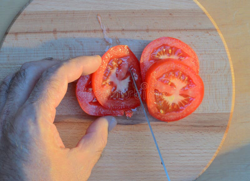Cortar un tomate con las manos con un cuchillo en una tabla de cortar de madera en la puesta del sol fotos de archivo libres de regalías