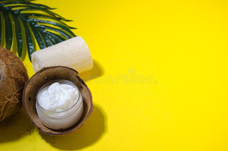 Cosmetic attributes, cream in a transparent jar and stands in a coconut, next to it is a loofah washcloth, a palm leaf on a yellow background. Shadows, soft focus, horizontal image